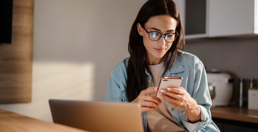 Serious charming woman using smartphone while working with laptop at home