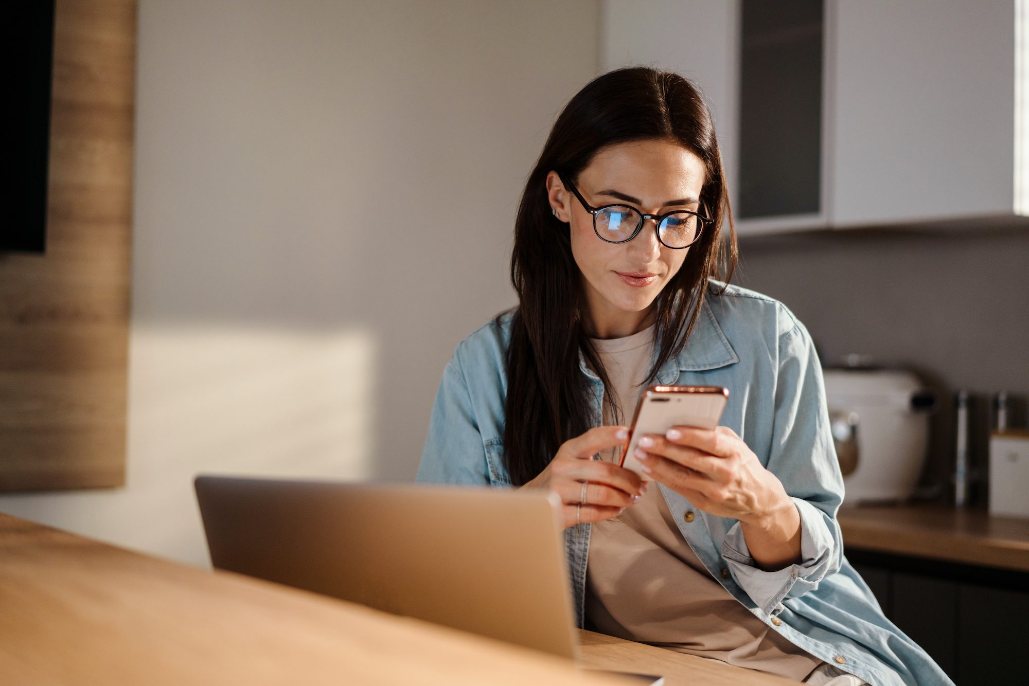 Serious charming woman using smartphone while working with laptop at home